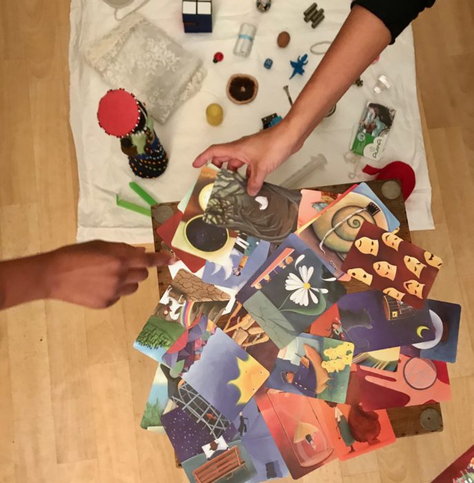 Zwei Hände greifen nach Bildkarten, daneben kleine Objekte auf einer Decke. 